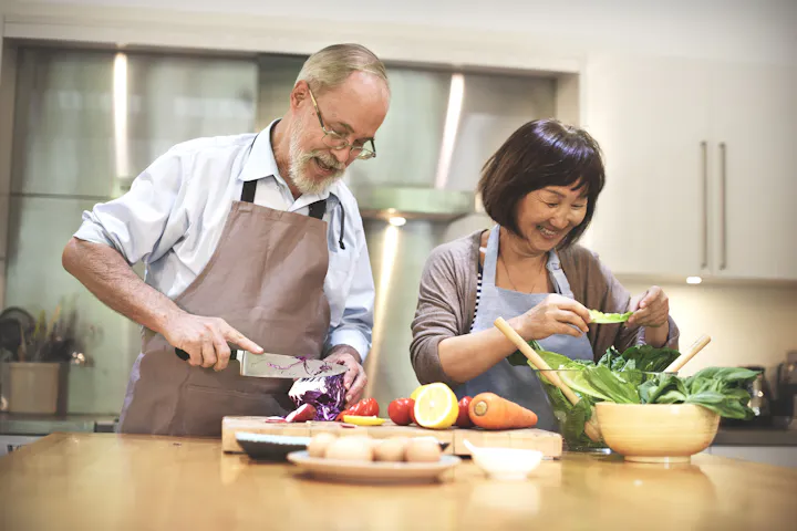 An older couple happily prepares a fresh, colorful meal together in a bright kitchen, surrounded by healthy vegetables and ingredients.