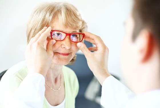 An older woman tries on a pair of red eyeglasses with the assistance of an eye care professional.
