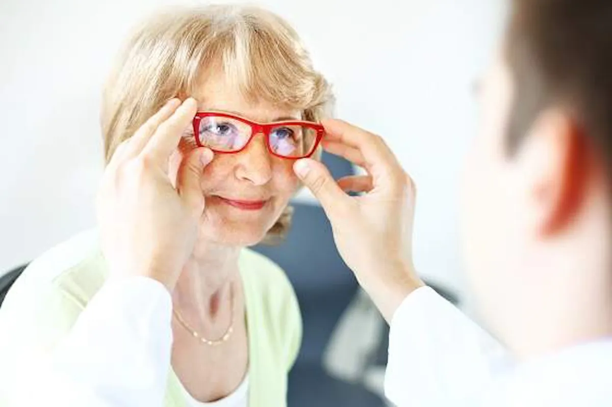 An older woman tries on a pair of red eyeglasses with the assistance of an eye care professional.