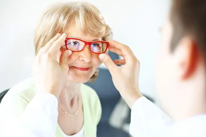 An older woman tries on a pair of red eyeglasses with the assistance of an eye care professional.