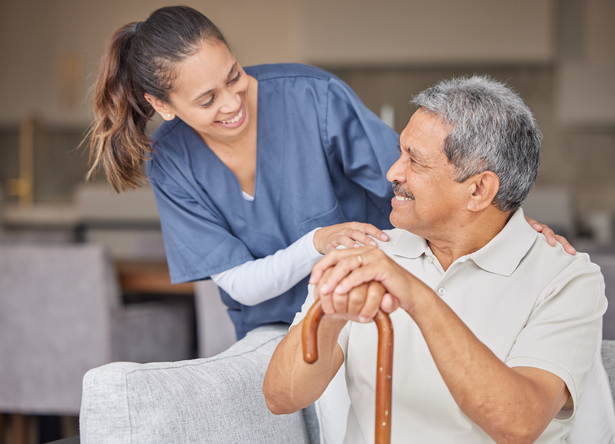 A caregiver smiles warmly while supporting an older man seated with a cane, reflecting companionship, care, and trust.