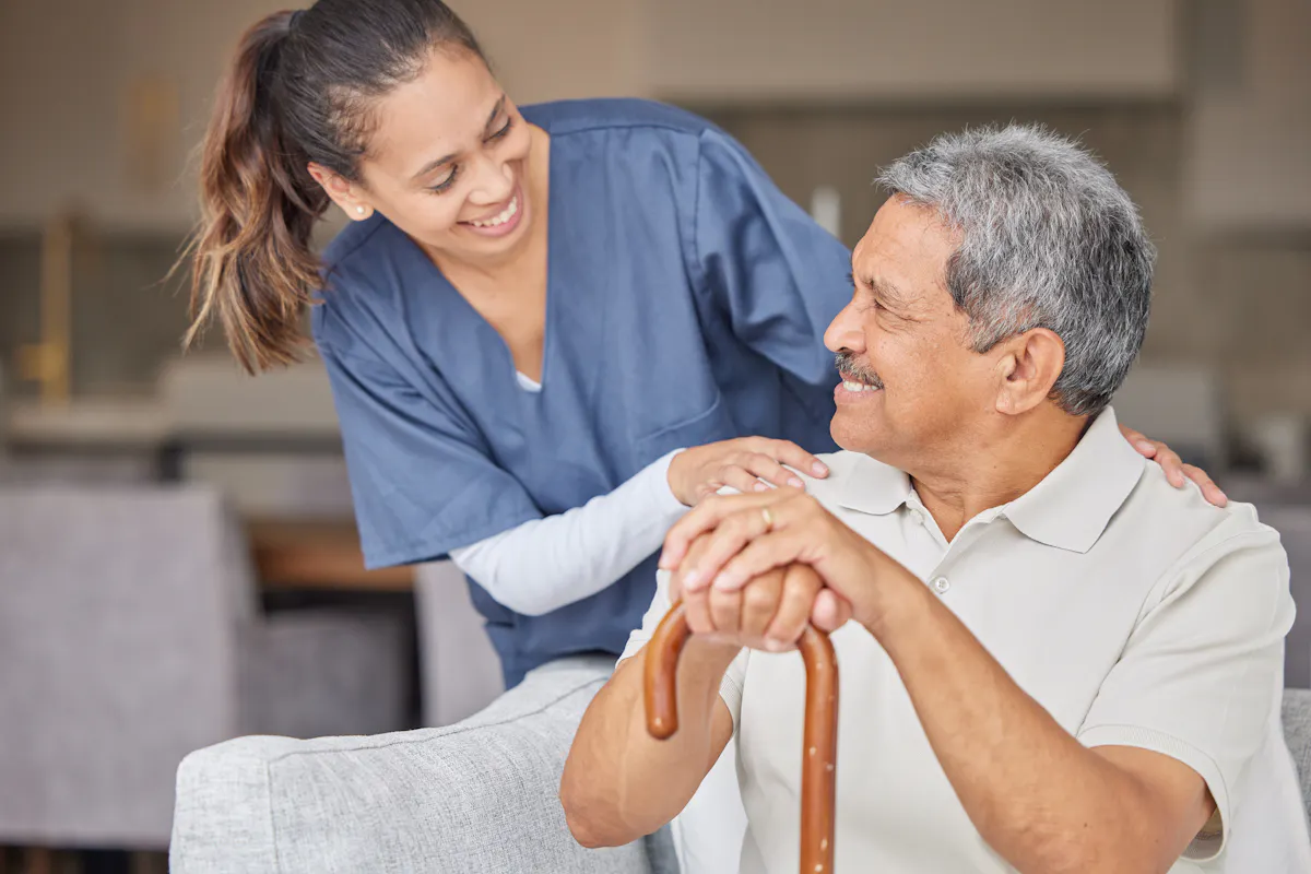 A caregiver smiles warmly while supporting an older man seated with a cane, reflecting companionship, care, and trust.