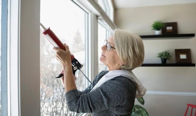 An older woman checks the seal around a window as part of preparing her home for colder winter weather.