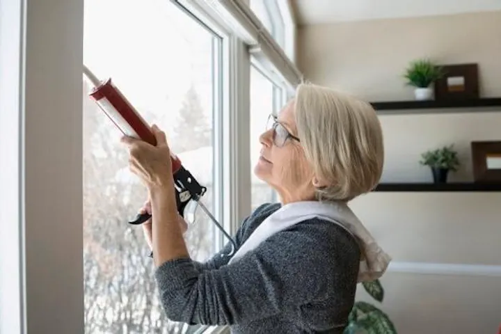 An older woman checks the seal around a window as part of preparing her home for colder winter weather.
