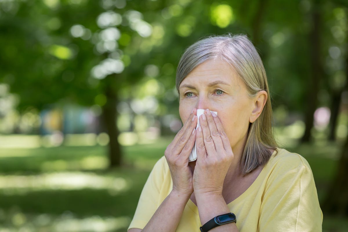An elderly woman in the park with allergies has a runny nose and sneezes near trees