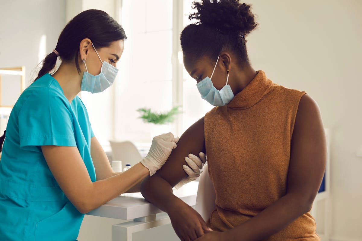 Young African American woman getting flu shot during seasonal vaccination campaign. Doctor or nurse in medical face mask cleans skin on patient's arm before injecting modern Covid 19 antiviral vaccine