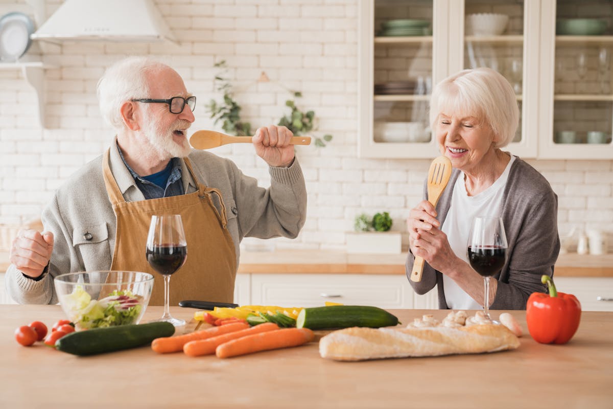 Love and relationship. Cheerful active seniors grandparents elderly spouses singing dancing together at home kitchen while cooking vegan vegetarian food meal, drinking wine, celebrating anniversary