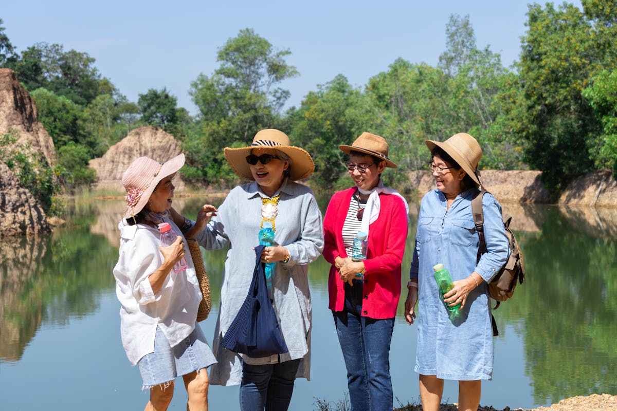 Group of retired elderly Asian women enjoy outdoor reunion traveling trip, casual clothing, smiling, holding water bottles, engaging in conversation, senior people surrounded by scenery sunny day