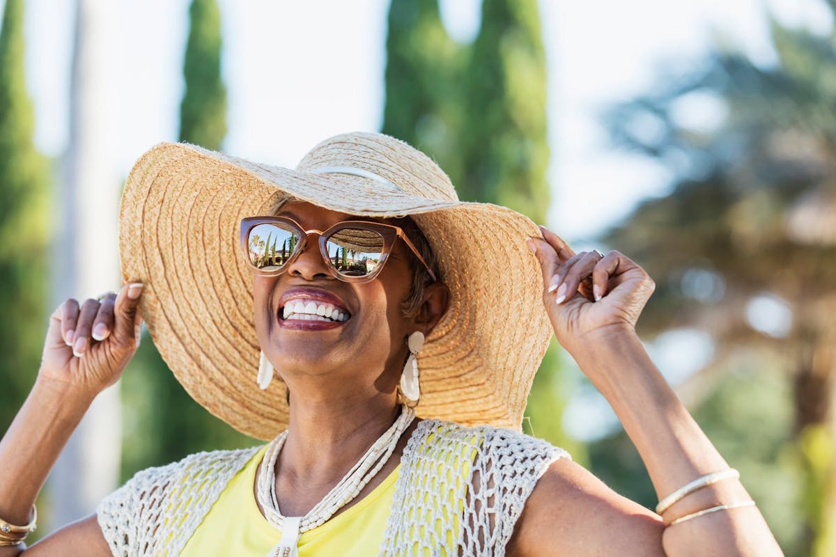 joyful woman enjoying a sunny day outdoors. She is smiling brightly while holding the brim of her wide straw sunhat. She’s wearing stylish sunglasses that reflect palm trees and the sky, giving the picture a vibrant, summery feel. Her outfit includes a yellow top, a light crochet-style vest, statement earrings, a layered necklace, and bangles