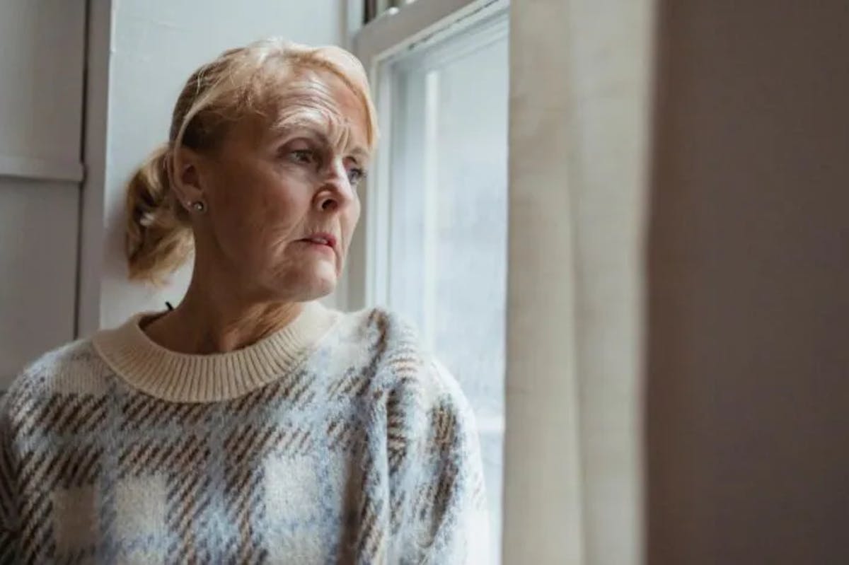elderly woman standing indoors near a window. She has light hair tied back and is wearing a warm, patterned sweater. Her expression appears thoughtful, reflective, or perhaps a bit sad as she gazes outside. The lighting is soft and natural, coming through the window, which gives the scene a quiet, contemplative mood.