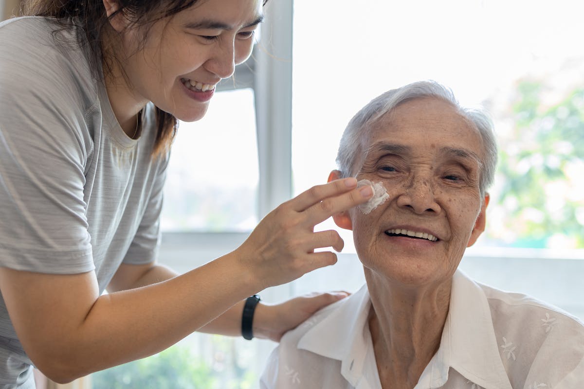 elderly woman smiling warmly while someone younger gently applies cream to her cheek