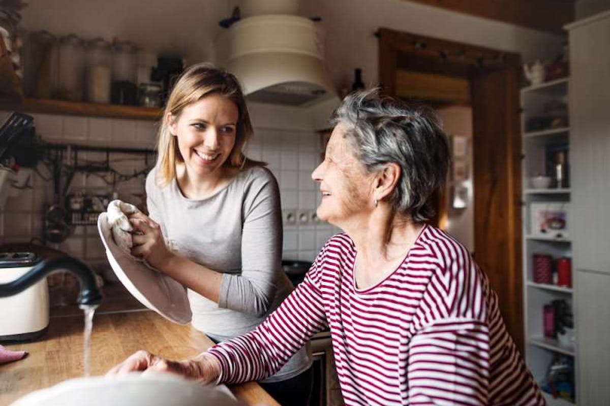 A younger woman and an older woman share a warm moment in the kitchen, smiling at each other while doing the dishes together.