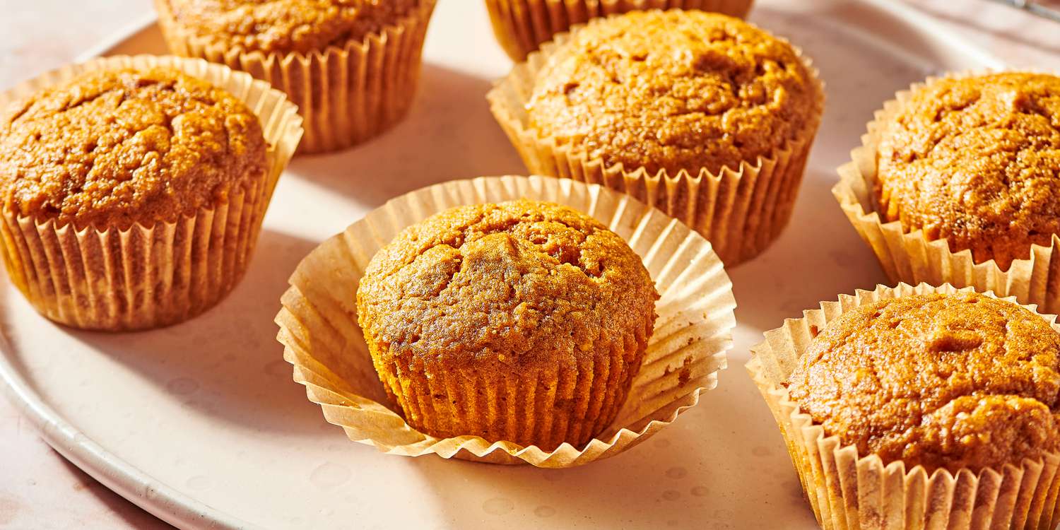Golden-brown muffins sit in crinkled paper liners, arranged closely together on a light-colored plate.