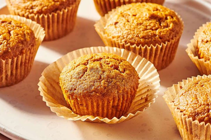 Golden-brown muffins sit in crinkled paper liners, arranged closely together on a light-colored plate.