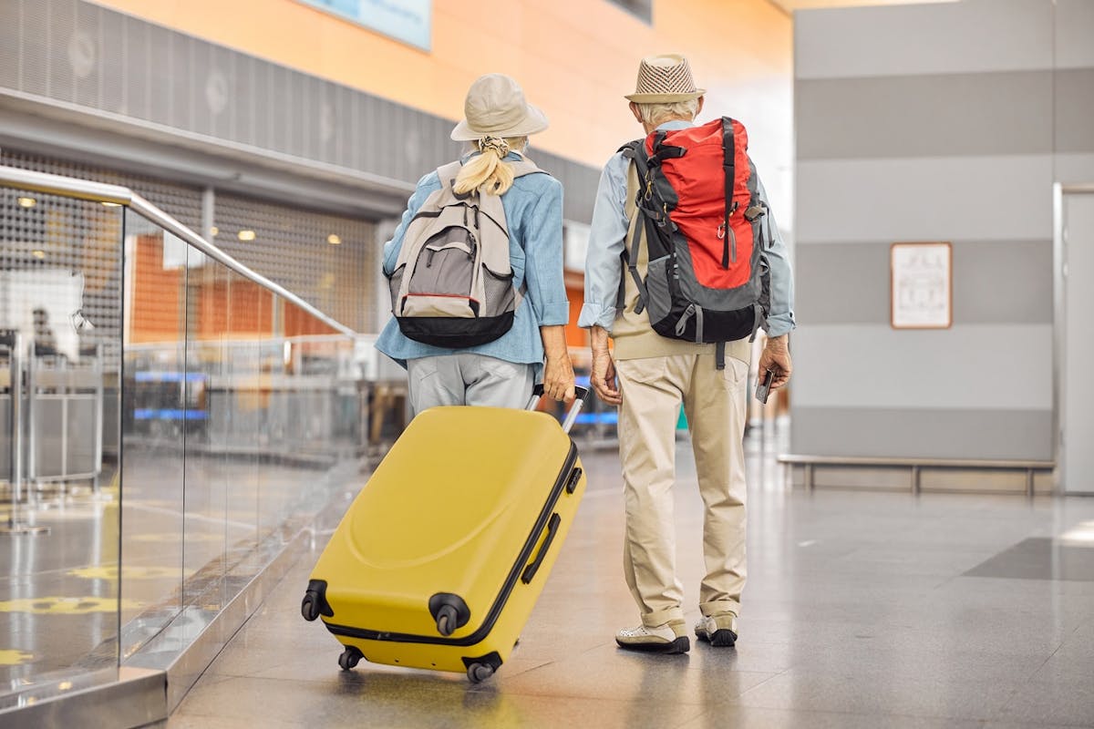 two older adults walking together through what appears to be an airport terminal. They are seen from behind, each wearing a backpack and sun hat, suggesting they are headed on a trip or vacation. One of them is pulling a bright yellow rolling suitcase, while the other holds a passport or travel document.
