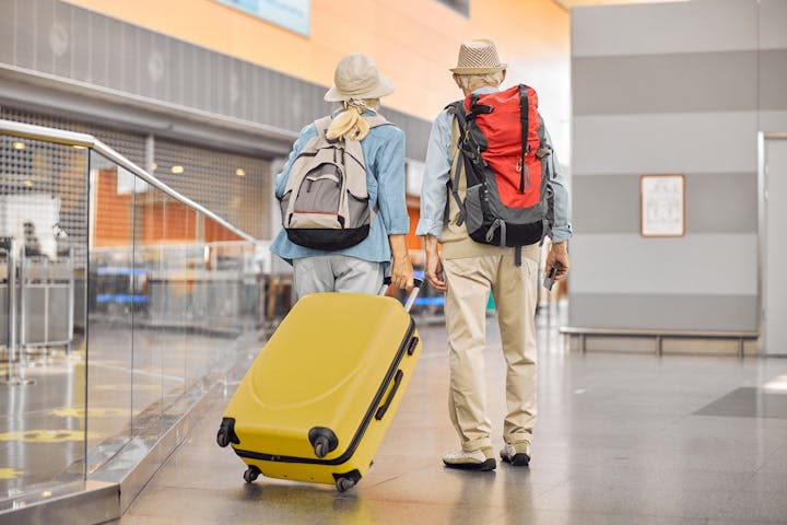 two older adults walking together through what appears to be an airport terminal. They are seen from behind, each wearing a backpack and sun hat, suggesting they are headed on a trip or vacation. One of them is pulling a bright yellow rolling suitcase, while the other holds a passport or travel document.