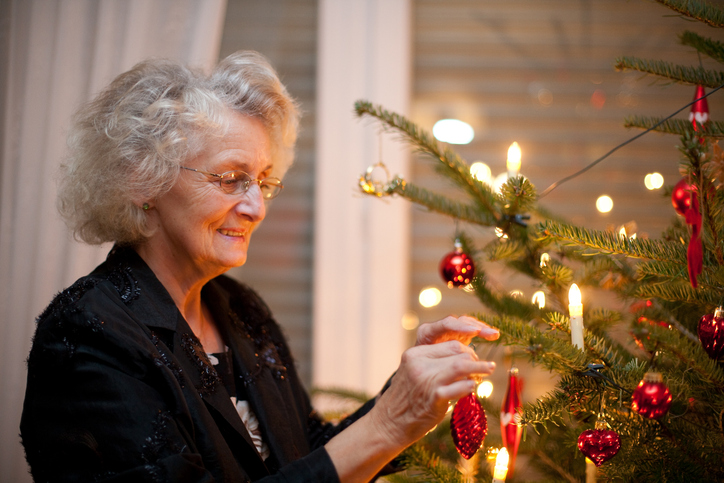 An older woman smiles warmly as she decorates a Christmas tree with ornaments and glowing candles.