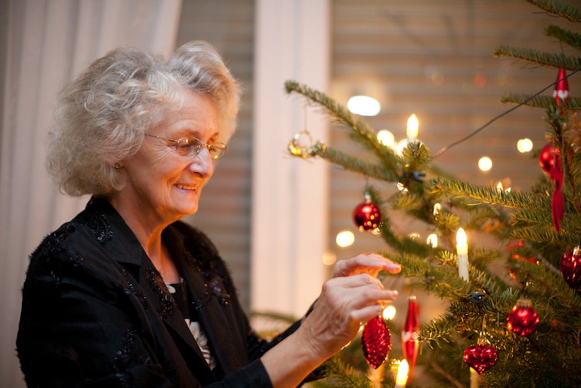 An older woman smiles warmly as she decorates a Christmas tree with ornaments and glowing candles.
