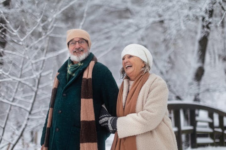 older couple walking outdoors in a snowy winter setting. They are bundled warmly in coats, scarves, and knit hats as they enjoy the cold weather. The woman is laughing joyfully while holding onto the man’s arm, and the man is smiling warmly. Behind them, snow-covered trees and a small wooden bridge create a peaceful, picturesque backdrop. The overall mood of the photo is happy, cozy, and full of companionship.