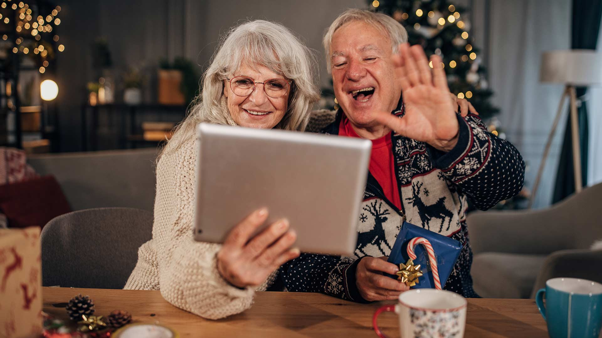 A smiling older couple sits at a festive holiday table, waving and chatting happily on a video call using a tablet.