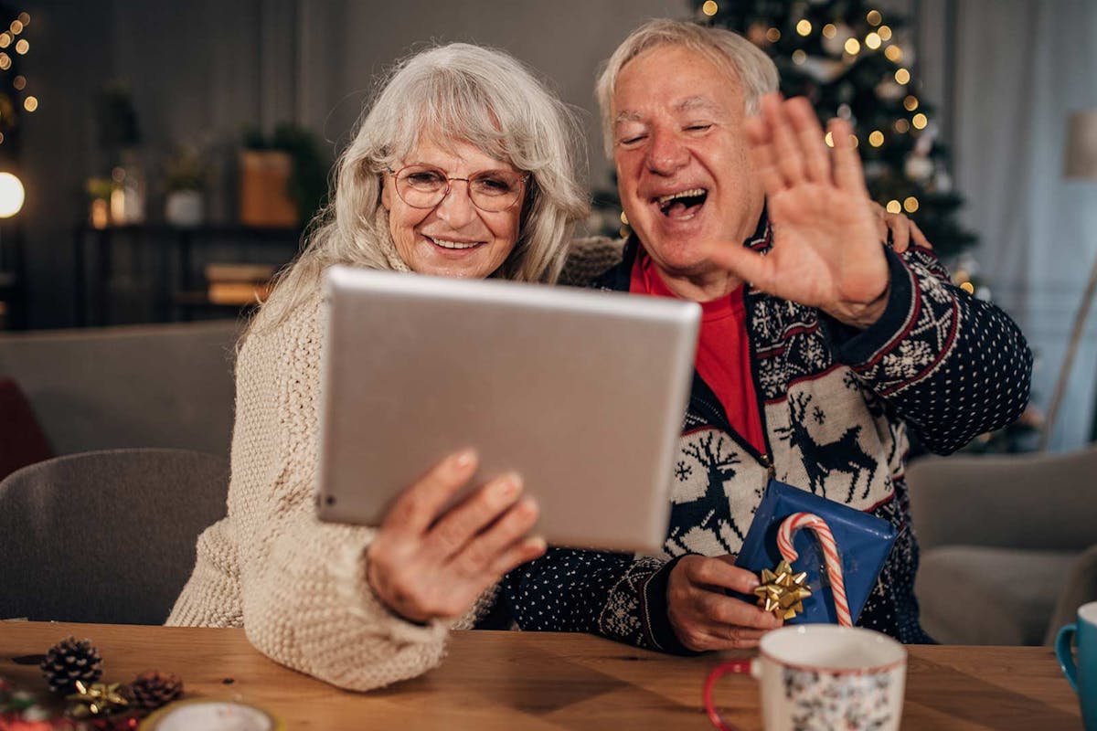 A smiling older couple sits at a festive holiday table, waving and chatting happily on a video call using a tablet.