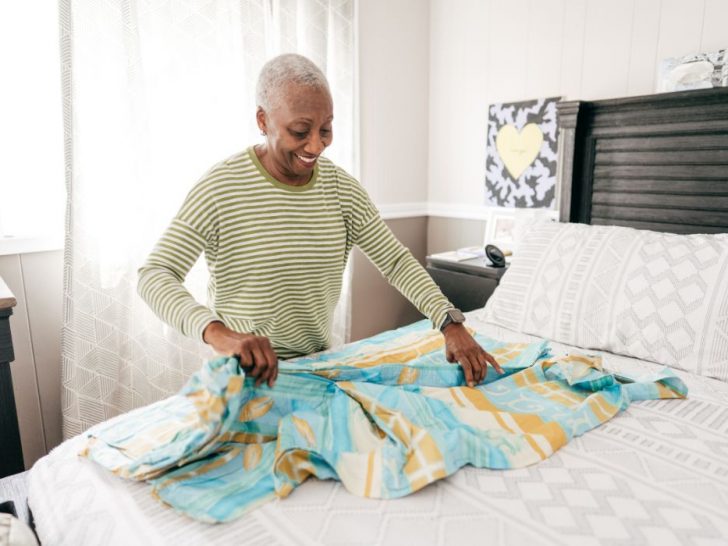 An older woman smiles as she neatly folds clothing on a bed in a bright, tidy bedroom, conveying a sense of calm, organization, and peaceful living.