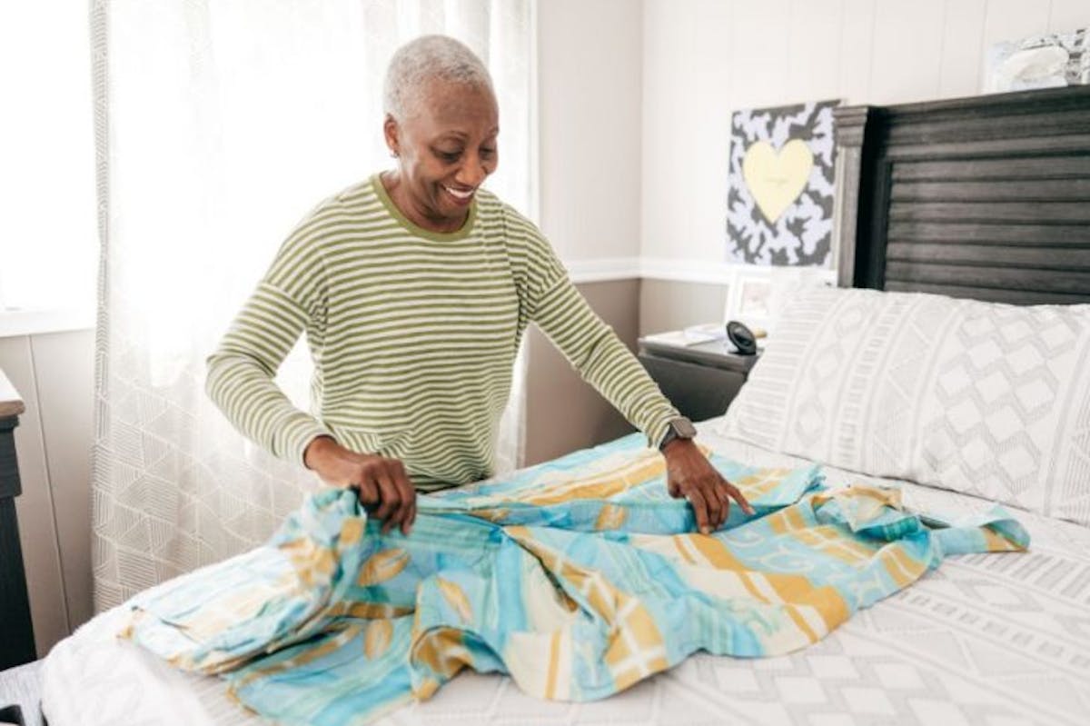 An older woman smiles as she neatly folds clothing on a bed in a bright, tidy bedroom, conveying a sense of calm, organization, and peaceful living.