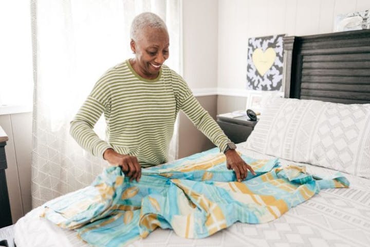 An older woman smiles as she neatly folds clothing on a bed in a bright, tidy bedroom, conveying a sense of calm, organization, and peaceful living.