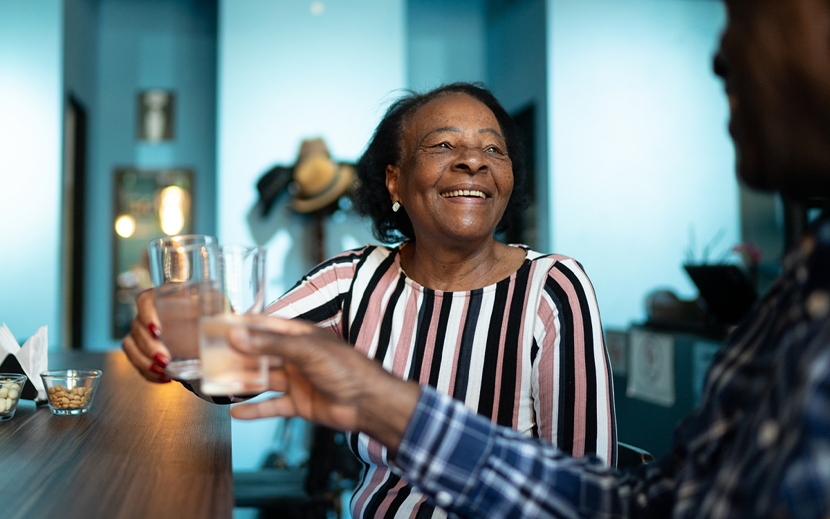 An older woman smiles warmly as she clinks glasses with another person at a table, sharing a relaxed and joyful moment together indoors.