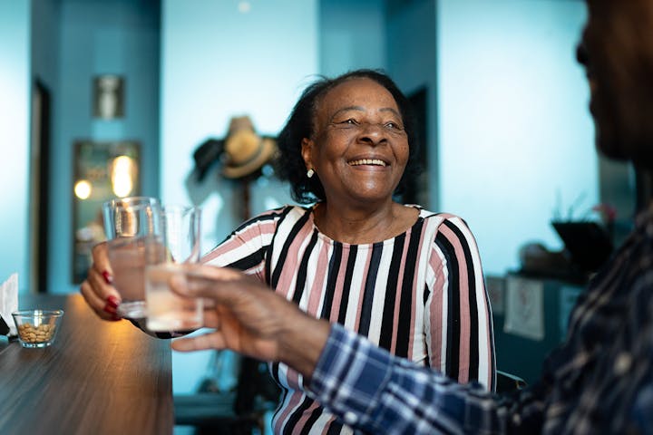 An older woman smiles warmly as she clinks glasses with another person at a table, sharing a relaxed and joyful moment together indoors.