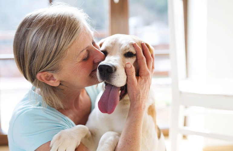 An older woman gently cuddles and kisses her happy dog, capturing a warm moment of love, companionship, and comfort between them.