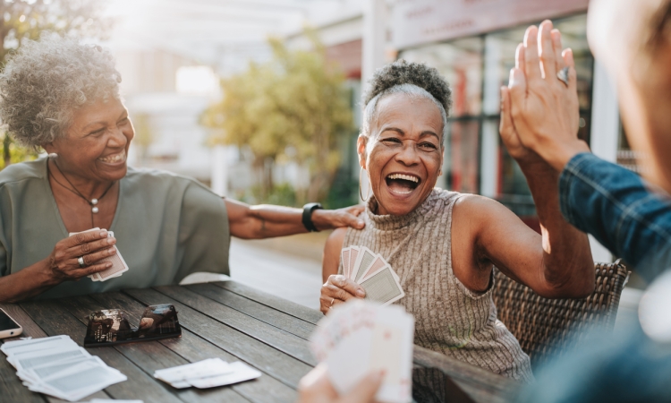 A group of older friends laugh and celebrate with a high-five while playing cards together outdoors, capturing the joy and connection of staying socially active.