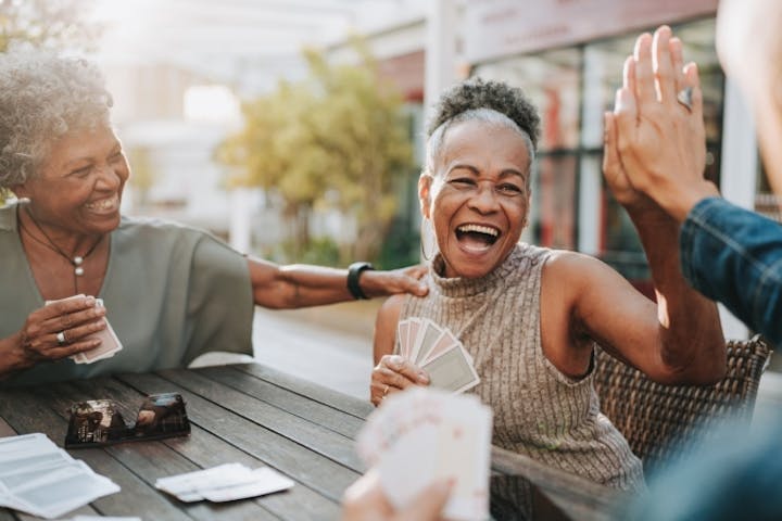 A group of older friends laugh and celebrate with a high-five while playing cards together outdoors, capturing the joy and connection of staying socially active.