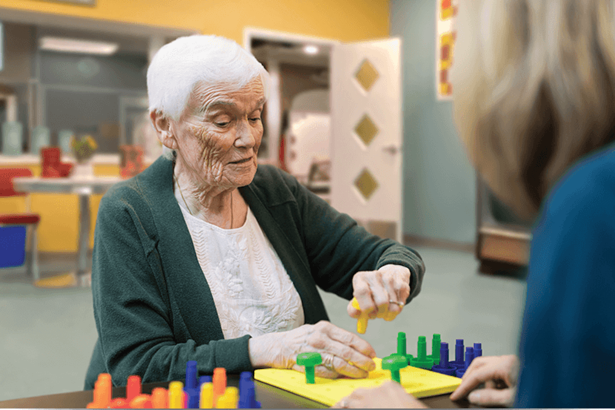 An elderly woman concentrates on placing colorful pegs into a puzzle at a table while a caregiver looks on in a bright, supportive activity space.
