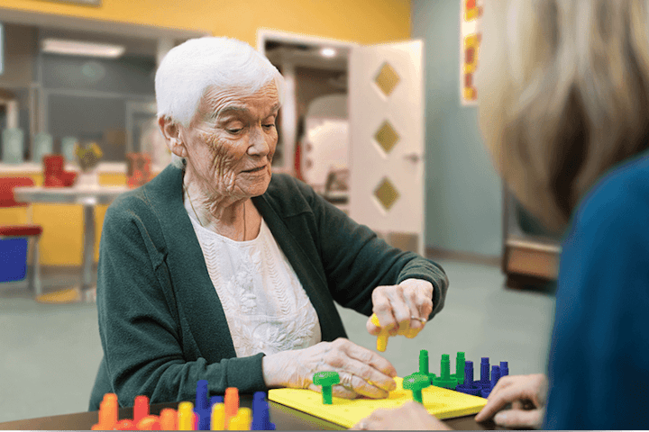 An elderly woman concentrates on placing colorful pegs into a puzzle at a table while a caregiver looks on in a bright, supportive activity space.