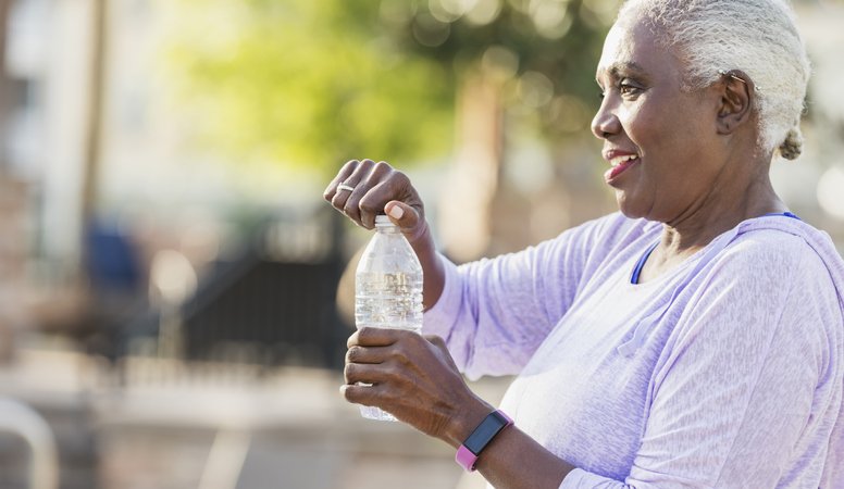 an older woman outdoors, smiling as she opens a bottle of water. She’s wearing a light purple long-sleeve top and a fitness tracker on her wrist. Her short gray hair and relaxed posture give the image a calm, healthy vibe.