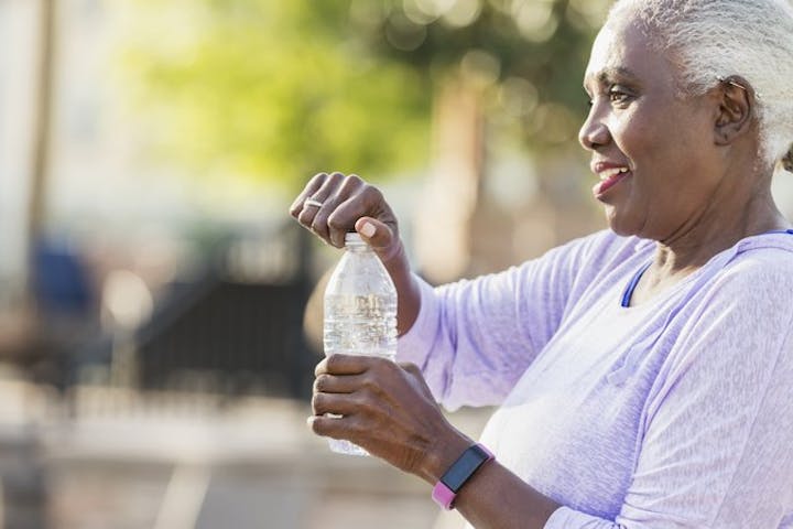 an older woman outdoors, smiling as she opens a bottle of water. She’s wearing a light purple long-sleeve top and a fitness tracker on her wrist. Her short gray hair and relaxed posture give the image a calm, healthy vibe.