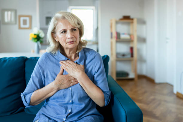 An older woman sitting on a couch holds her chest with a concerned expression, suggesting she may be experiencing chest discomfort or pain.