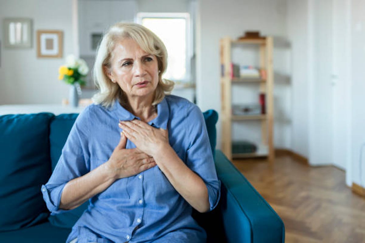 An older woman sitting on a couch holds her chest with a concerned expression, suggesting she may be experiencing chest discomfort or pain.
