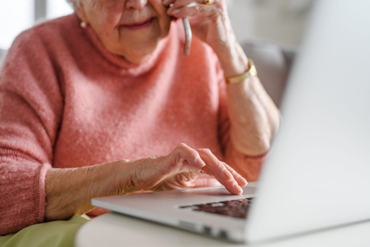 An elderly woman in a pink sweater is using a laptop while talking on the phone, suggesting she may be handling an online matter or call.