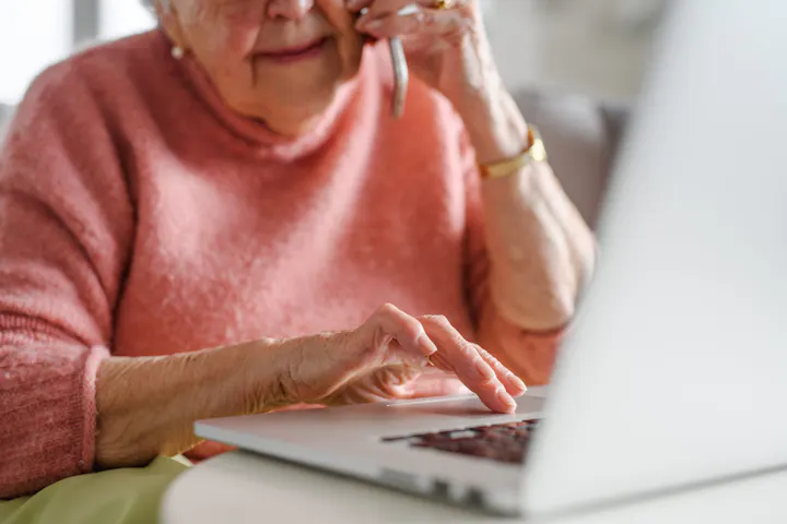 An elderly woman in a pink sweater is using a laptop while talking on the phone, suggesting she may be handling an online matter or call.