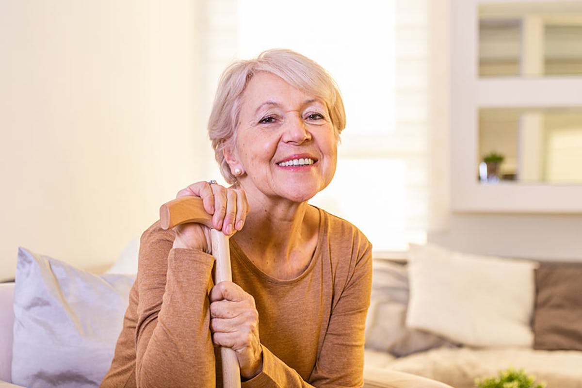 A smiling older woman holding a cane sits comfortably in a bright, cozy living room, conveying warmth, independence, and confidence.