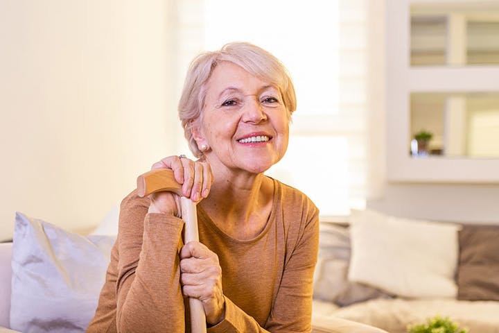 A smiling older woman holding a cane sits comfortably in a bright, cozy living room, conveying warmth, independence, and confidence.