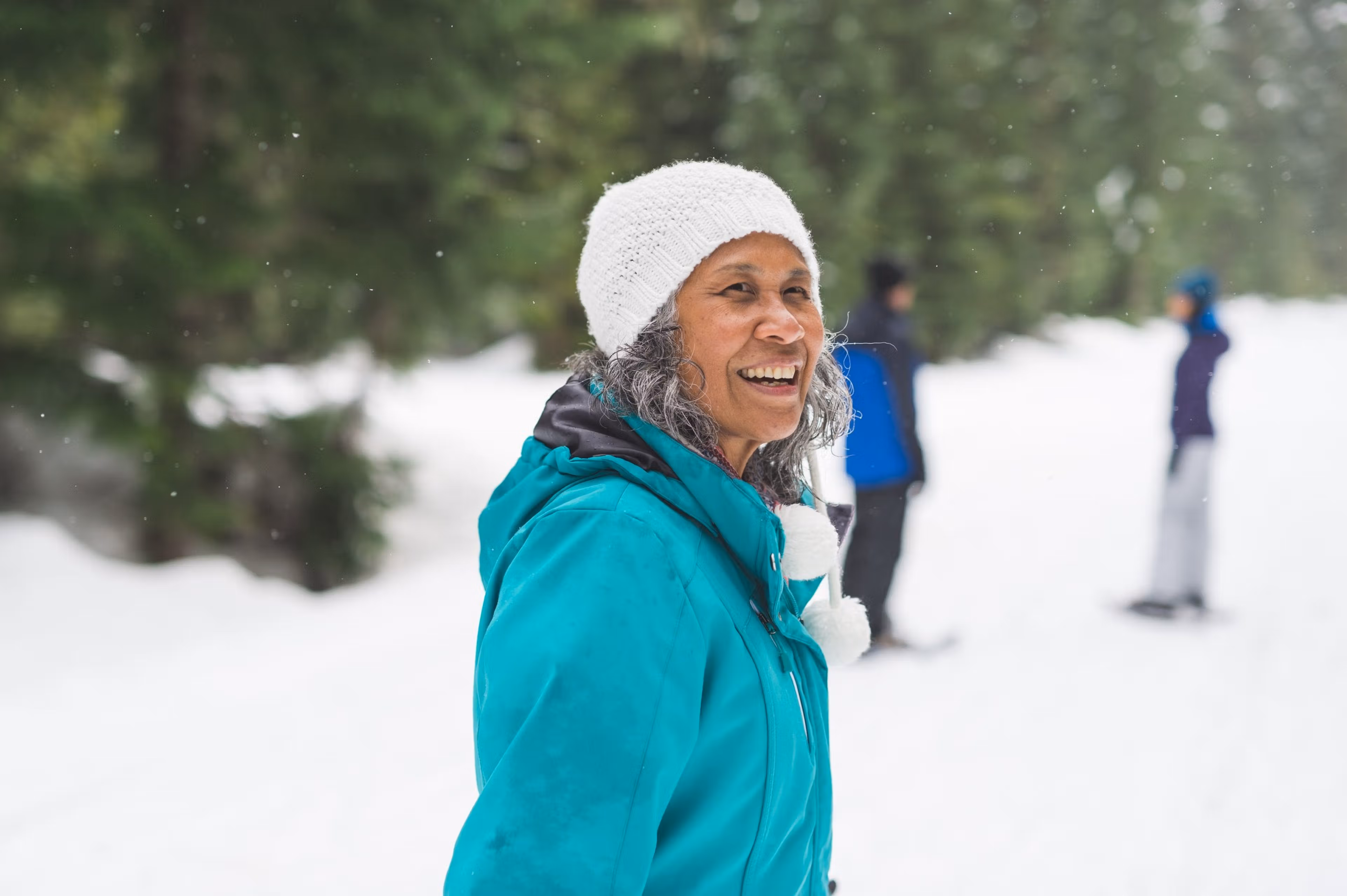 A smiling older woman in a teal winter jacket and white knit hat stands outdoors in a snowy forest while other people are blurred in the background.
