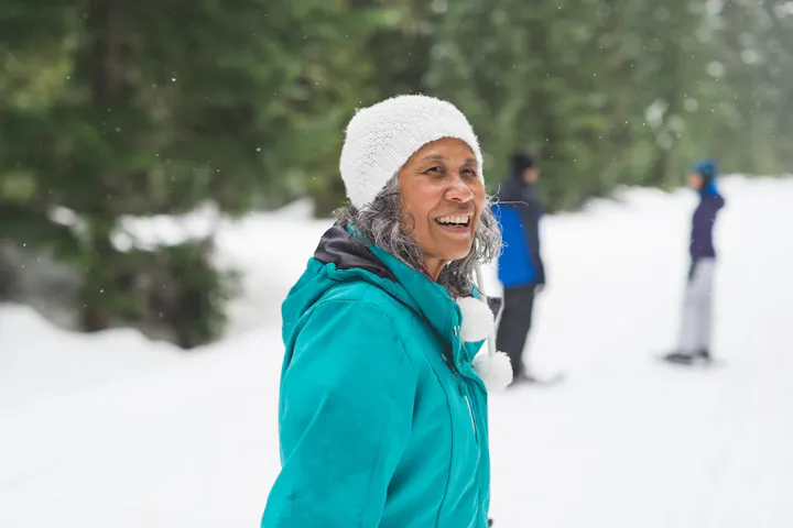 A smiling older woman in a teal winter jacket and white knit hat stands outdoors in a snowy forest while other people are blurred in the background.