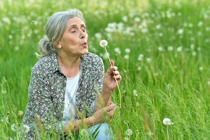 An elderly woman sits in a grassy field, gently blowing on a dandelion as its seeds drift into the air.