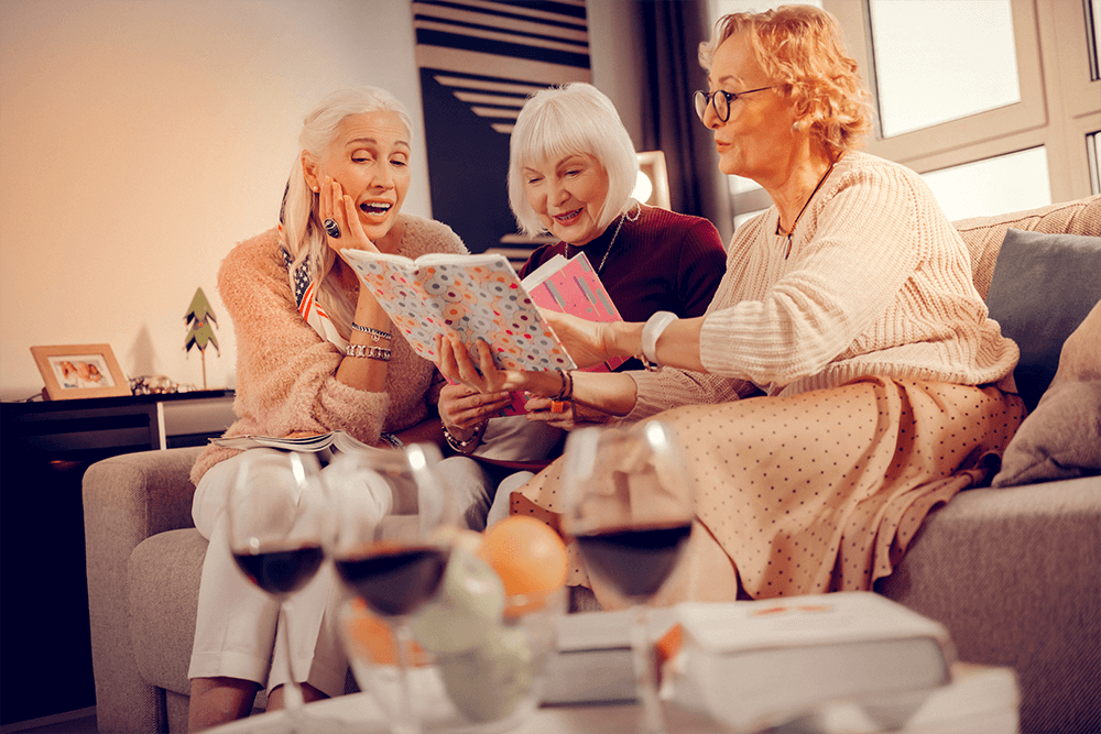 Three older women sit together on a couch, smiling and chatting as they look through a book, with glasses of wine on the table in front of them.