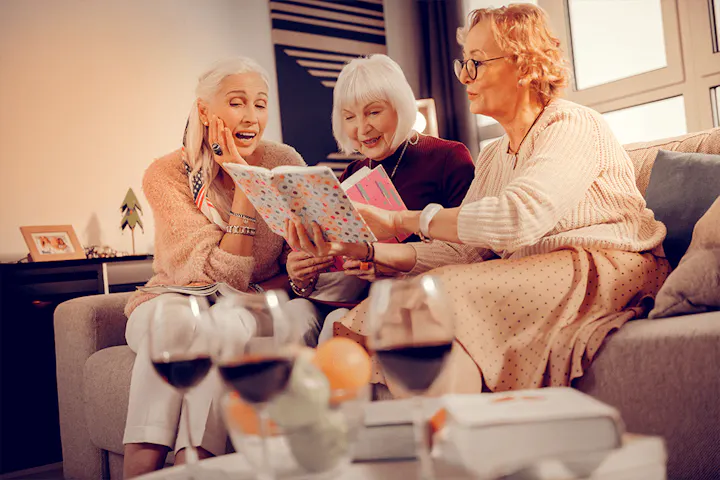 Three older women sit together on a couch, smiling and chatting as they look through a book, with glasses of wine on the table in front of them.
