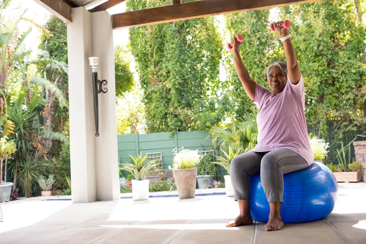 An older woman sits on a blue exercise ball on a covered patio, smiling as she lifts small pink dumbbells overhead during a gentle outdoor workout.