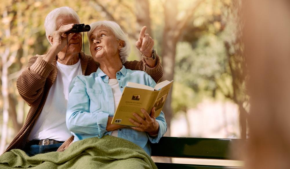 An elderly couple sits on a park bench in autumn, with the man looking through binoculars while the woman points into the distance and holds an open book.